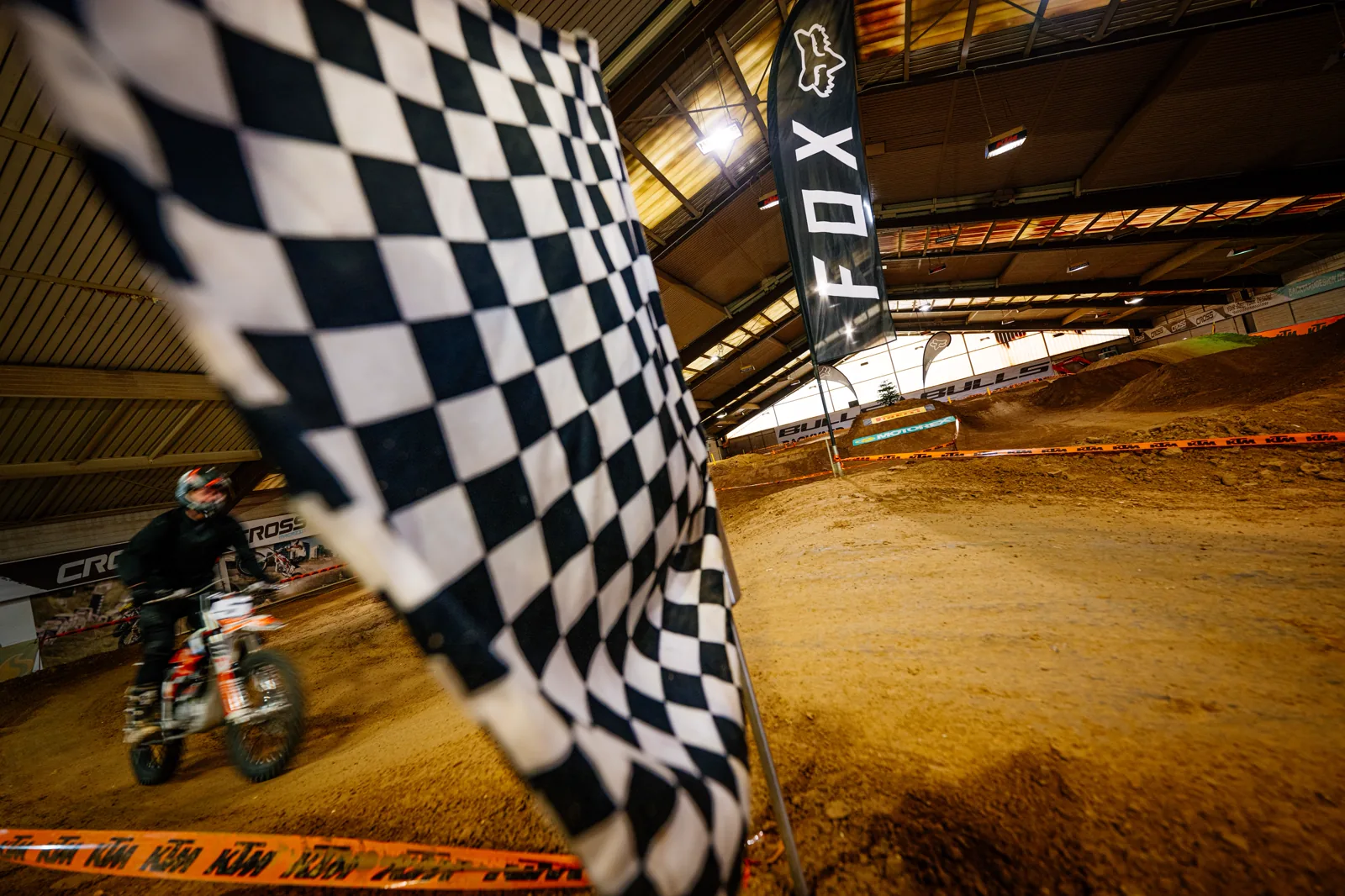Schwarz-weiße Zielflagge in der Indoor-Halle der eDirtarena, dahinter ein Fahrer auf der Strecke